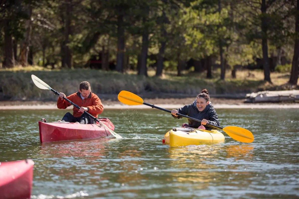 This image features two kayakers, each in a brightly colored kayak, paddling on a shimmering river surrounded by lush green trees. The water's surface reflects the sky, and the scene radiates a sense of adventure and connection to nature.