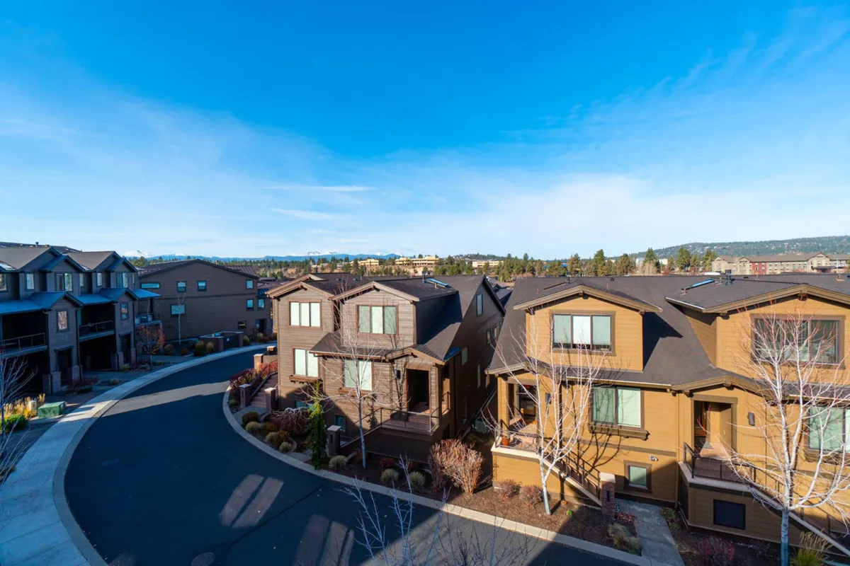 An exterior view of residential buildings lining a curved road, surrounded by landscaped gardens and outdoor spaces. The image captures people walking and socializing within a friendly, open neighborhood setting, hinting at access to trails and communal outdoor zones.