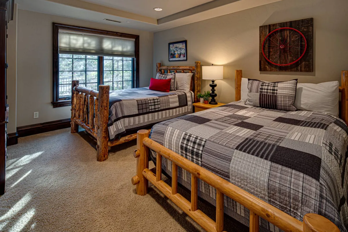 A cozy bedroom with log-style twin beds dressed in plaid bedding, a bedside lamp, and a window view. The room invites relaxation and togetherness, offering a warm, rustic feel ideal for families or groups.