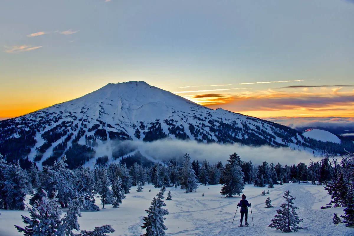 A breathtaking view of a snow-capped mountain glowing in the setting sun, bordered by snow-covered trees. In the foreground, a skier is visible, adding a sense of adventure and connection to the vibrant outdoor lifestyle Bend is known for.