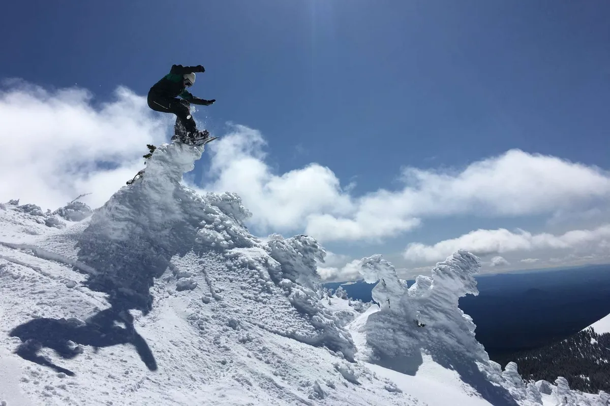 A snowboarder is caught mid-action on a snow-covered mountain peak beneath a bright blue sky, with panoramic views of the surrounding snowy landscape. The image conveys excitement, energy, and the thrill of winter sports.