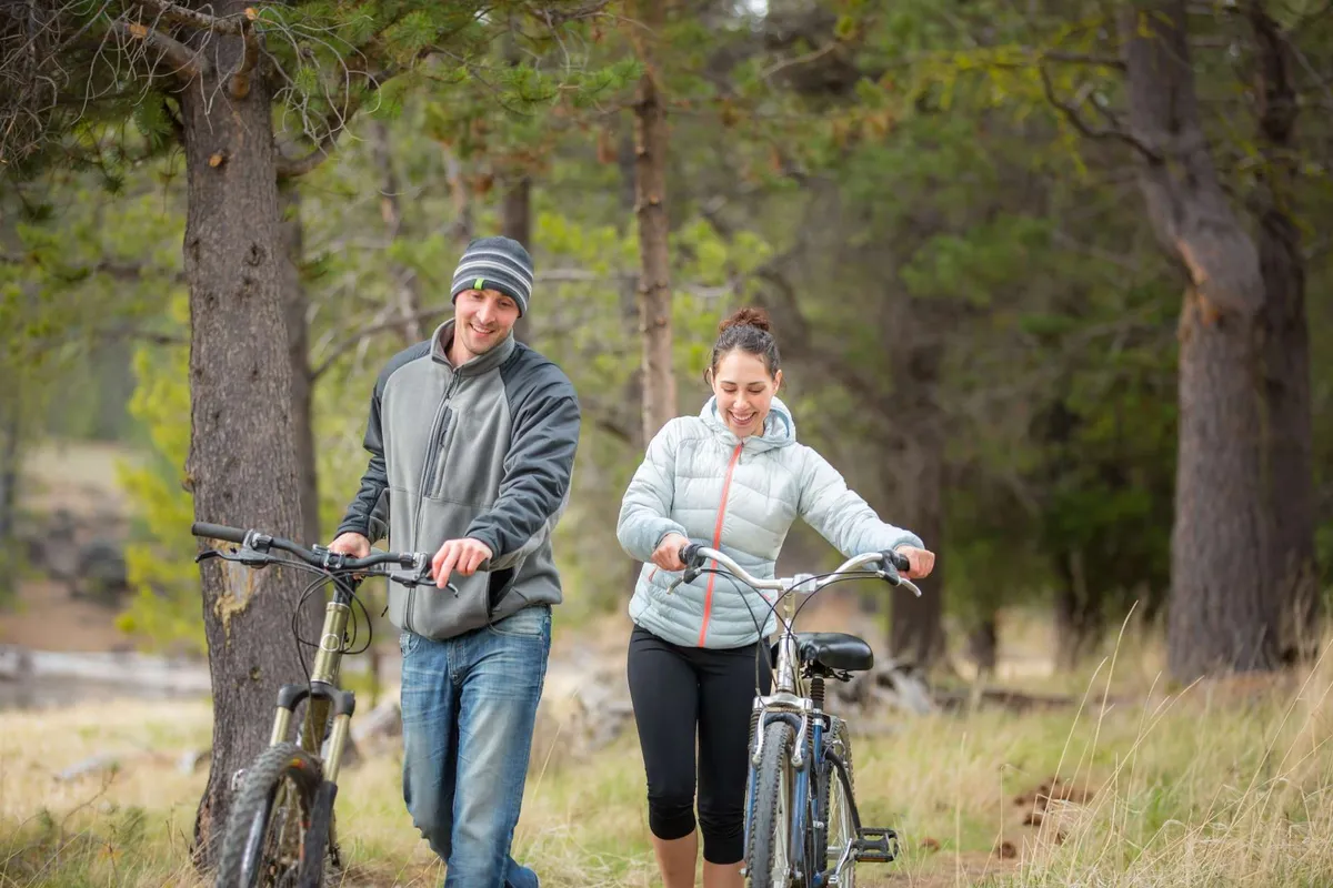 Two bicycles are parked on a path while a couple strolls nearby, surrounded by tall trees and greenery. The atmosphere is inviting, suggesting exploration and shared outdoor experiences in a scenic environment.