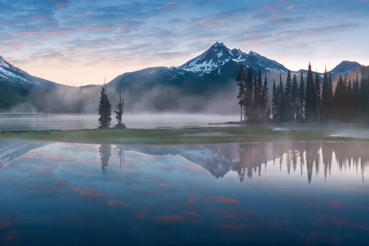 This image showcases a tranquil mountain and lake view, framed by trees and an expansive sky. The landscape evokes the natural beauty of Bend, Oregon, highlighting opportunities for outdoor exploration alongside your dog.