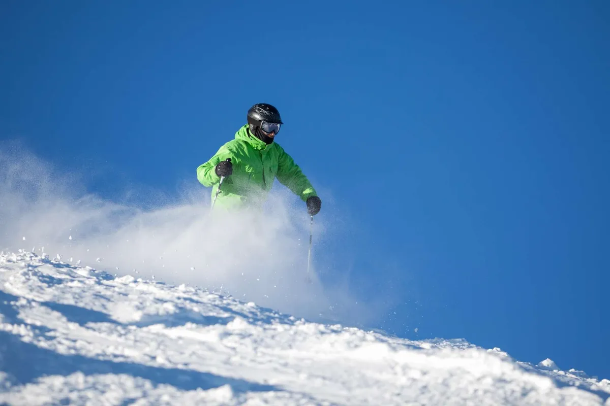 An action-packed exterior shot of a skier in a vibrant green jacket gliding down a snow-covered slope beneath a blue sky. The surroundings capture the excitement and activity potential near Deschutes Crest.