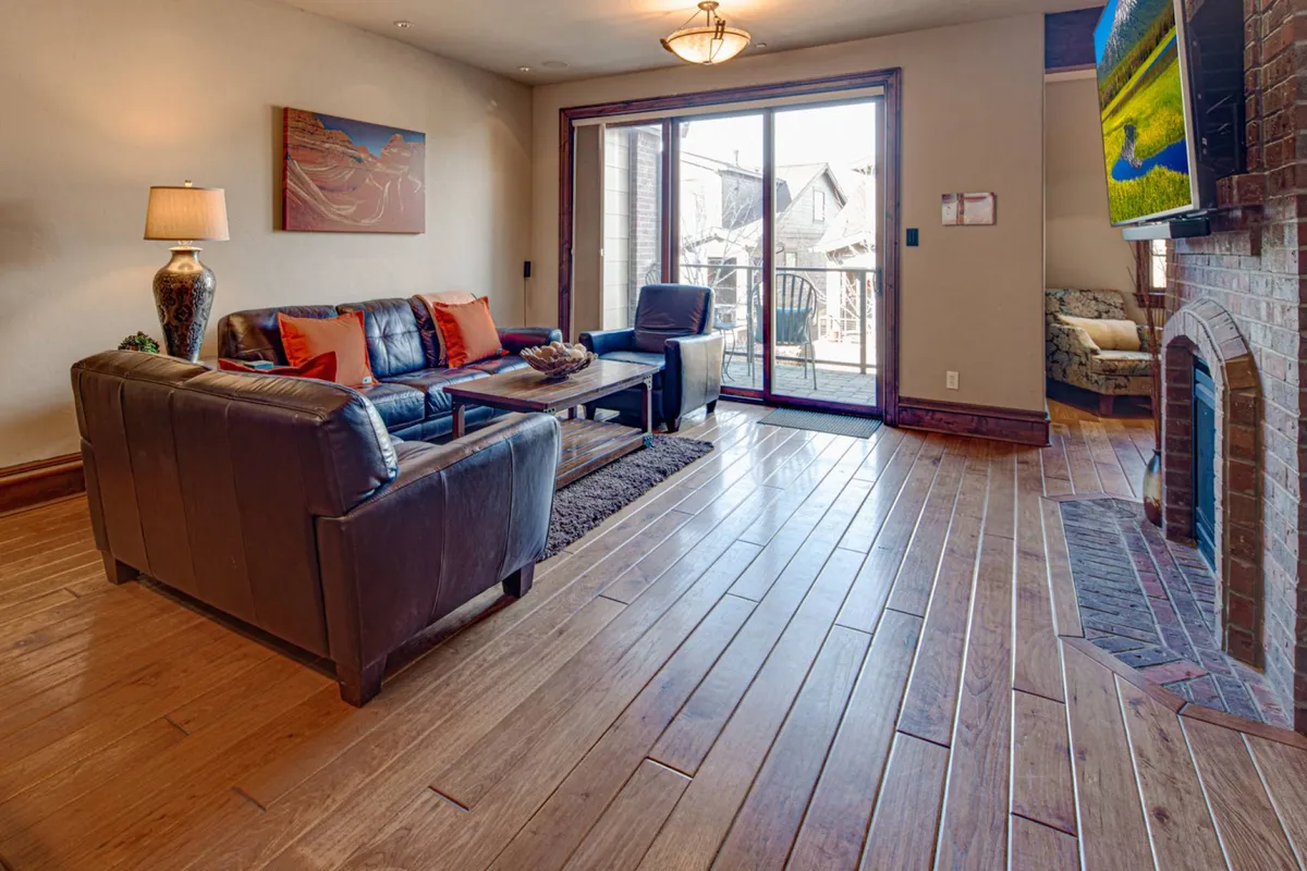 This image captures a cozy living area featuring a rich leather sofa, coffee table, and a striking fireplace. A large sliding glass door opens to the outside, flooding the room with natural light and offering a glimpse of the views beyond. Artwork on the wall adds a layer of warmth and sophistication, while the setup invites relaxation and lively conversation.