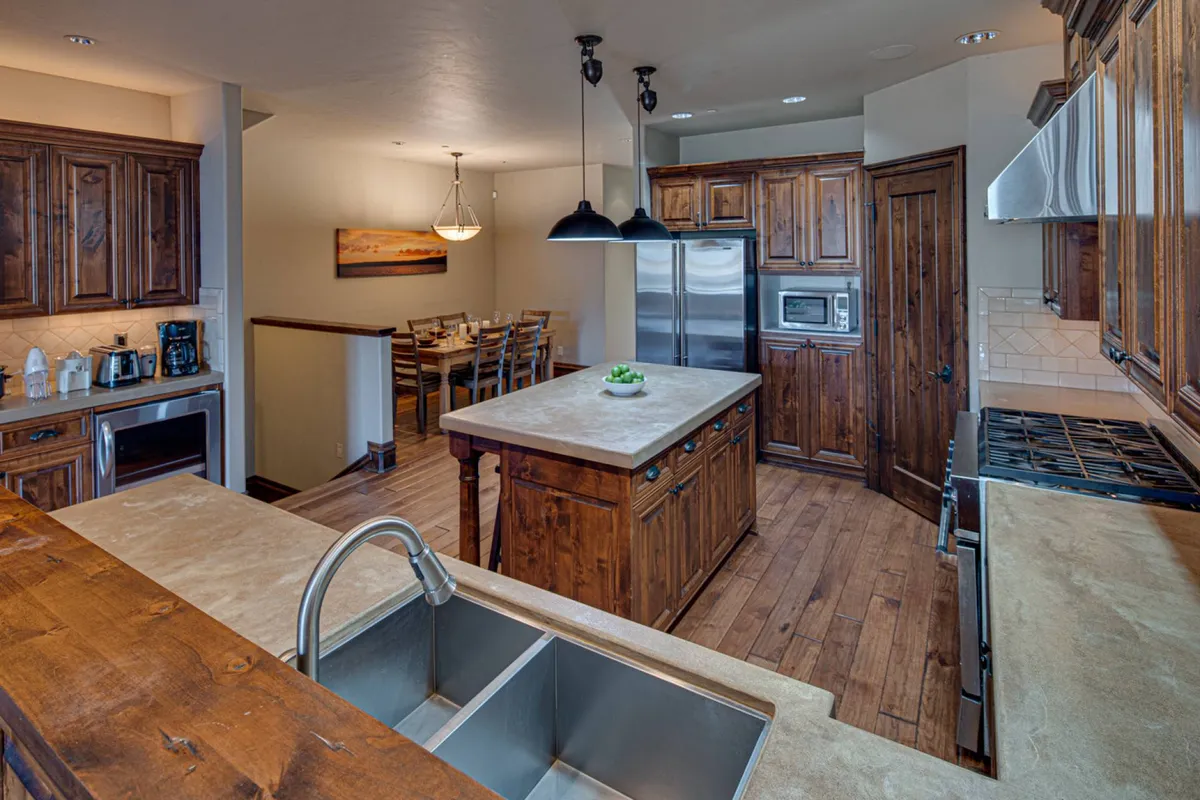 This image showcases an open-concept kitchen with a central island featuring a sink, surrounded by warm wooden cabinetry. In the background, a dining area is visible, suggesting space for gatherings and culinary experiences. The inviting atmosphere highlights the kitchen's role as a focal point for entertaining and group activities.