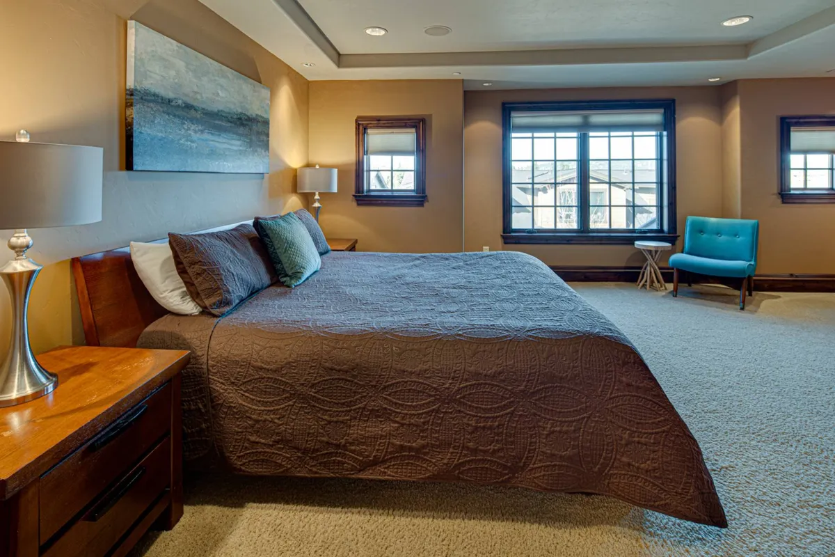 This image showcases a serene bedroom with a king-sized bed, an art piece above the headboard, and a blue accent chair in the corner. Natural light streams in, creating a tranquil, restful atmosphere ideal for relaxation or quiet moments.