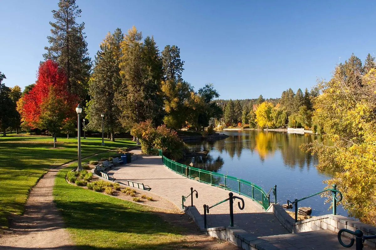 A tranquil exterior scene featuring a calm lake edged by colorful trees and a walking path with green railings. Park benches are visible, inviting visitors to sit and enjoy the natural beauty. The scene captures a peaceful, walkable area where people can relax, picnic, or explore nature just steps from the property.
