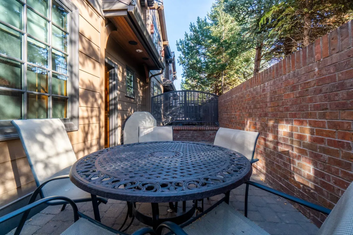 A cozy garden patio featuring a round table surrounded by greenery and a brick wall. The setup is inviting for outdoor dining, with seating ready for an al fresco meal beneath the open sky—perfect for enjoying BBQs or starlit evenings.