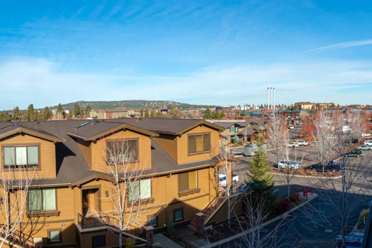 A welcoming exterior shot showcasing modern architecture surrounded by tall trees beneath a clear blue sky. The view hints at spacious outdoor areas perfect for relaxing, with visible mountain vistas in the distance. The setting feels alive, with the sense of community, neighborhood exploration, and local events subtly suggested by the inviting layout and environment.